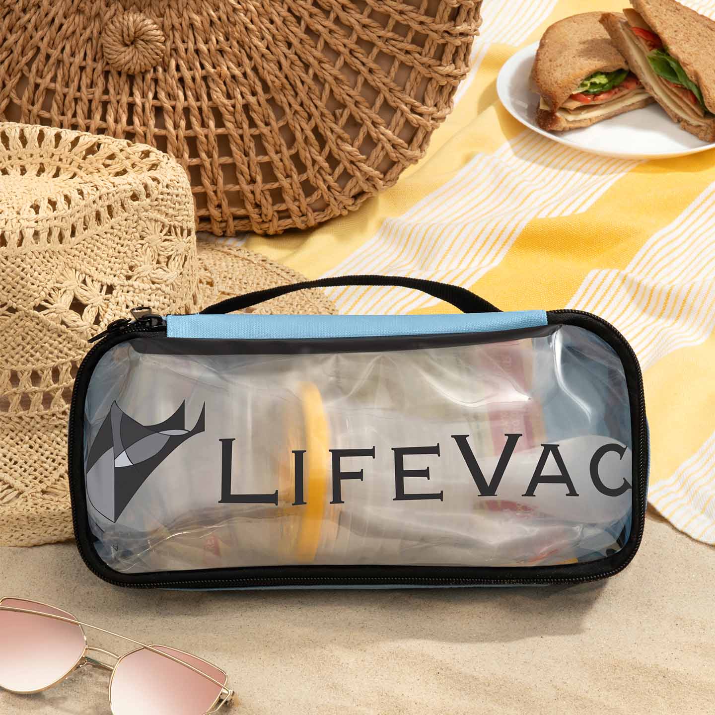 Blue LifeVac Travel Kit on the sand next to beach blanket, hat, sunglasses, bag, and sandwich